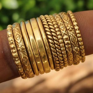 Pode incluir: Uma coleção de anéis dourados, alguns com padrões intrincados, empilhados em um dedo. Os anéis variam em design, incluindo faixas lisas, texturas trançadas e formas geométricas. Fundo verde desfocado.