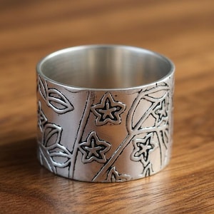 May include: A silver-colored ring with a floral and star pattern. The ring is cylindrical with a detailed design of leaves and five-pointed stars etched around the band. The ring sits on a wooden surface.