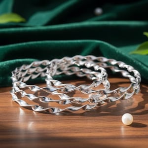 May include: Three silver-colored, twisted-design bangles are displayed on a wooden surface, with a dark green velvet backdrop. The bangles are the focal point, reflecting light. Scattered pearls and green leaves add to the aesthetic.