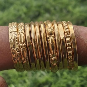 May include: A collection of gold-colored bangles, some with intricate floral and geometric patterns, stacked on a finger. The bangles vary in design, with some featuring smooth surfaces and others with textured details. The background is a blurred green.