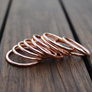 Könnte beinhalten: Eine Sammlung dünner roségoldener Ringe auf einer dunklen Holzoberfläche. Die Ringe sind glatt und glänzend und reflektieren das Licht. Die Ringe sind übereinander gestapelt, wodurch ein geschichteter Effekt entsteht.
