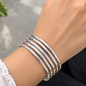 May include: A set of five polished silver-toned bangles worn together on the wrist. The bracelets feature a smooth, rounded, and reflective surface. Each bangle is identical in shape and size, creating a stacked appearance on the arm.