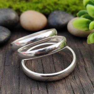 May include: Three silver rings are interlocked, forming a distinctive design. The rings have a smooth, reflective surface with a polished finish. They are presented on a dark wooden surface, with a blurred background of greenery and stones.