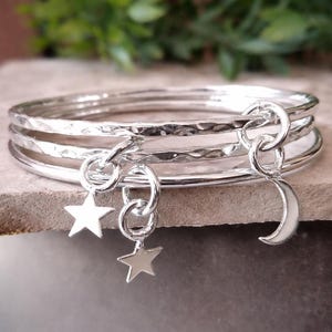 Puede incluir: Un conjunto de cuatro pulseras plateadas con textura martillada. Dos colgantes de estrella y un colgante de luna creciente cuelgan de las pulseras. Las joyas se exhiben sobre una superficie neutra con follaje verde de fondo.