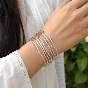 Op de afbeelding: Een set van zes zilverkleurige armbanden, elk met een opvallend golvend en golvend ontwerp. Deze metalen armbanden worden samen als een stapel gedragen, wat een gelaagd uiterlijk creëert met hun gladde, reflecterende oppervlakken en consistente gebogen vormen.