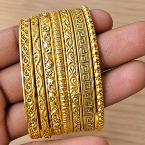 Puede incluir: Un conjunto de siete brazaletes de metal color oro con distintos patrones en relieve, incluyendo formas geométricas, líneas onduladas, motivos florales, diseños de hojas repetidas y un patrón de greca, con un borde de cuentas.