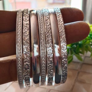 May include: A collection of silver bangles, some with floral and dot patterns, others smooth. The bangles are stacked together, reflecting light. The jewelry is likely for fashion or adornment.