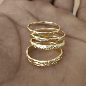 May include: A stack of four gold-colored metal rings. The top two rings feature a smooth, scalloped edge design. The third ring has a delicate engraved floral pattern, and the bottom ring displays a repeating geometric cross-like motif.