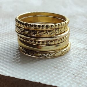 Peut inclure: Une pile de cinq bagues dorées aux textures variées. L'ensemble comprend deux anneaux à bords perlés, un anneau à surface lisse et polie, un anneau avec un motif floral gravé et un anneau avec un motif en croix finement ciselé.