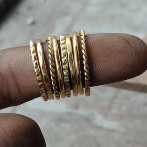 May include: A set of seven thin gold-toned rings featuring various textures, including smooth, twisted rope-like patterns, and indented geometric designs. These stackable rings are arranged side-by-side, showcasing a polished metallic finish.