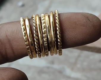 May include: A set of seven thin gold-toned rings featuring various textures, including smooth, twisted rope-like patterns, and indented geometric designs. These stackable rings are arranged side-by-side, showcasing a polished metallic finish.