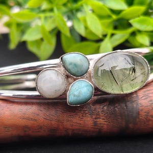 May include: A silver-toned cuff bracelet featuring an array of gemstones. The bracelet has a central cluster of stones, including a large oval green stone, two blue stones, and a white stone. The bracelet is displayed on a wooden surface with green foliage in the background.