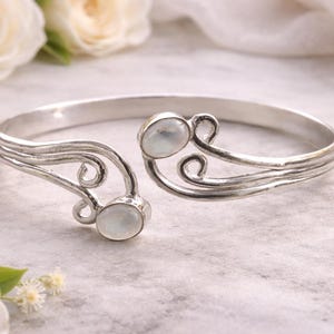 Puede incluir: Una pulsera de puño plateada con dos piedras ovaladas de color claro. La pulsera presenta un diseño fluido y texturizado con remolinos decorativos. El diseño abierto permite un uso fácil. La pulsera se muestra sobre una superficie marmórea.