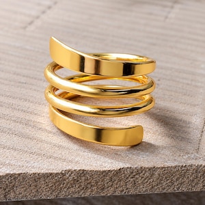 May include: A gold-toned ring featuring a multi-layered spiral design. The band consists of a continuous metal coil that wraps around itself, creating four distinct horizontal loops with a smooth, polished, and reflective surface finish.