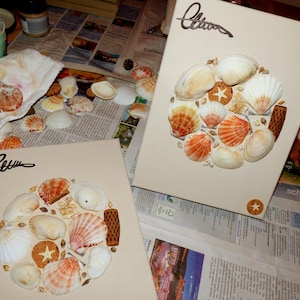 Könnte beinhalten: Zwei beige Leinwände mit einer kreisförmigen Anordnung von Muscheln, Seesternen und dekorativen Elementen. Die Muscheln sind weiß und orange, mit einer Signatur oben links. Die Leinwände liegen auf einer mit Zeitungspapier bedeckten Oberfläche.