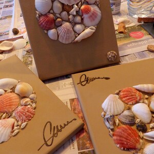 Puede incluir: Tres lienzos marrones decorados con una disposición circular de conchas marinas. Las conchas son de varios tamaños y colores, incluyendo blanco, rosa y naranja. Cada lienzo tiene una firma manuscrita en tinta marrón.