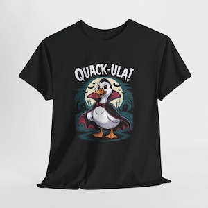 Op de afbeelding: Zwart T-shirt met een cartoon eend verkleed als vampier, compleet met cape en vlinderdas. De tekst "QUACK-ULA!" staat boven de eend. De achtergrond toont een volle maan, bomen en grafstenen.