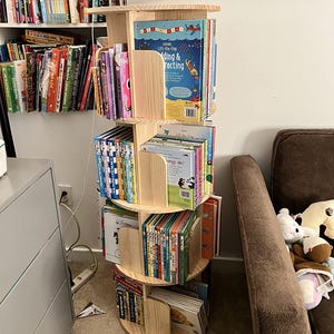 May include: A wooden, rotating bookshelf filled with colorful children's books. The bookshelf has multiple tiers and a circular design, allowing easy access to the books. The books are of various sizes and colors, with some titles visible.