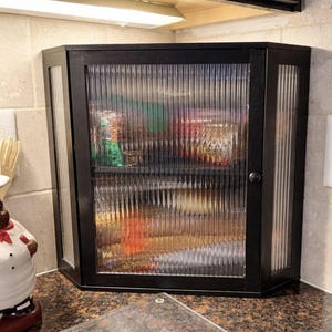 May include: A black corner cabinet with a ribbed glass door and side panels. The cabinet is mounted on a countertop and appears to be in a kitchen. The cabinet is closed and the contents are not visible.