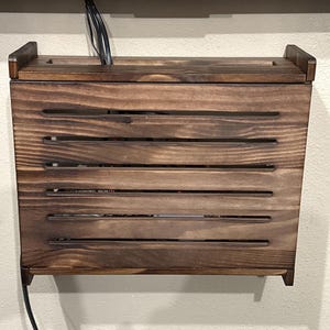 May include: A stained wooden media box with horizontal slats and a top lid. The box is mounted on a wall and has a cable running from the bottom. The box is designed to hold media devices.