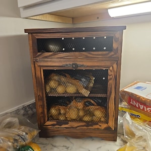 May include: A wooden onion storage cabinet with a dark stain. The cabinet has a mesh front and contains several bags of onions. The cabinet is sitting on a countertop with other produce nearby.