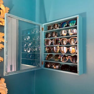 May include: A blue wall-mounted sunglasses display cabinet with a mirrored door. The cabinet is open, revealing multiple pairs of sunglasses on shelves. The sunglasses have various frame and lens styles.