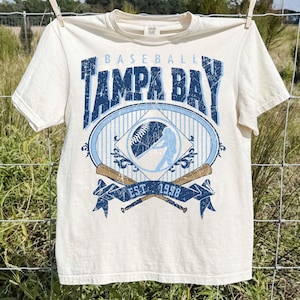 May include: Cream-colored baseball t-shirt with a distressed blue graphic. The graphic reads "Tampa Bay Baseball" with a baseball and player silhouette. Below the graphic are crossed bats and the text "EST. 1998".