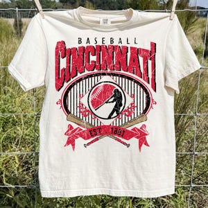 May include: Cream-colored baseball t-shirt featuring the word "Cincinnati" in distressed red lettering. The shirt includes a baseball graphic with a batter silhouette, crossed bats, and the text "EST. 1881".