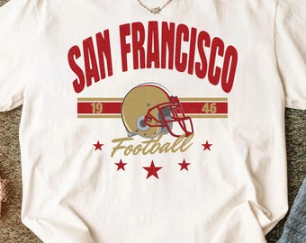 Può includere: Una t-shirt bianca a maniche corte con una grafica rossa che recita SAN FRANCISCO sopra un casco da football americano dorato con maschera rossa. Una striscia rossa orizzontale contiene i numeri 19 e 46, con la scritta Football e cinque stelle.