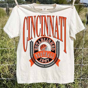 Könnte beinhalten: Ein cremefarbenes Kurzarm-T-Shirt mit einer großen orangefarbenen und schwarzen Grafik. Der Text lautet oben CINCINNATI, darunter ESTABLISHED FOOTBALL 1967, umgeben von einem Football-Helm-Symbol und flankiert von zwei vertikalen Blattmotiven.