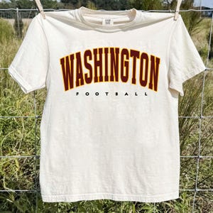 May include: A cream-coloured short-sleeve t-shirt featuring the text WASHINGTON in a large, arched, dark red font with a yellow outline, and the word FOOTBALL in a smaller, black, sans-serif font centred directly underneath the main text.