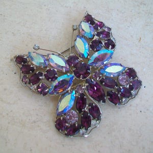 May include: A butterfly-shaped brooch with a silver-tone metal frame. The brooch is adorned with various sizes of purple and iridescent oval and round rhinestones. The wings and body are detailed with the sparkling stones.