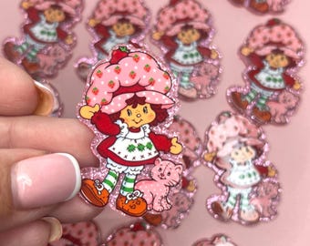 May include: A collection of Strawberry Shortcake character stickers. Each sticker features the character in a red dress and strawberry hat, with a pink cat. The stickers are on a pink background.