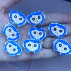 May include: A collection of eight blue and white shoe charms featuring a cartoon character with blue hair, button eyes, and a small red bow. The charms are designed to be attached to shoes and are held in a hand.