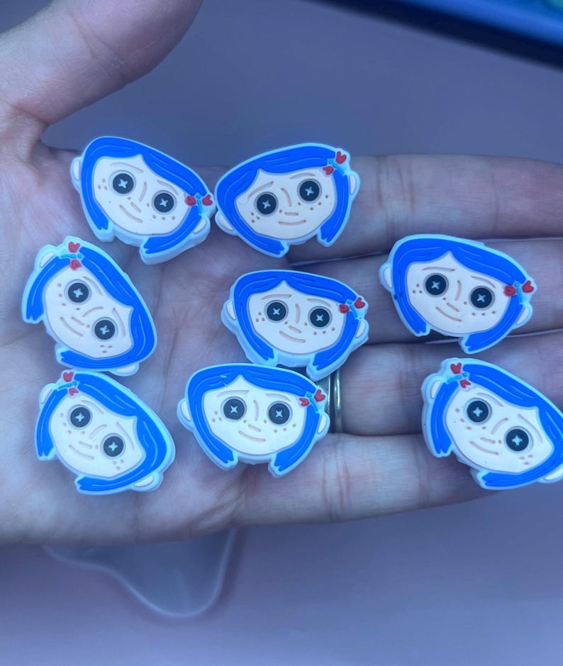 May include: A collection of eight blue and white shoe charms featuring a cartoon character with blue hair, button eyes, and a small red bow. The charms are designed to be attached to shoes and are held in a hand.