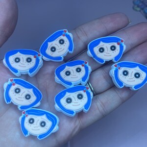 May include: A collection of cartoon character charms featuring a girl with blue hair, black button eyes, and a pale complexion. Each charm has a small red heart detail. The charms are likely for shoes or accessories.