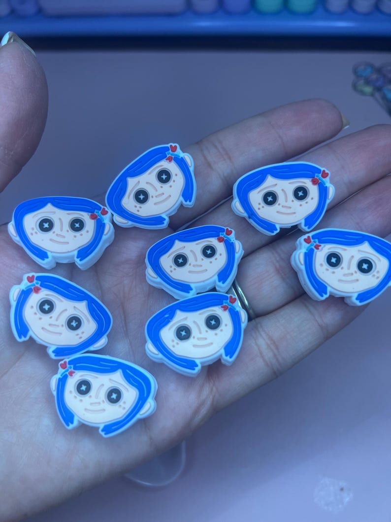 May include: A collection of cartoon character charms featuring a girl with blue hair, black button eyes, and a pale complexion. Each charm has a small red heart detail. The charms are likely for shoes or accessories.
