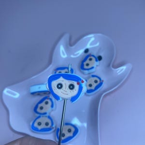 May include: A collection of blue and white character-themed charms and a decorative pick. The charms feature a cartoon face with blue hair and a red bow. The pick is held in a pink dish.