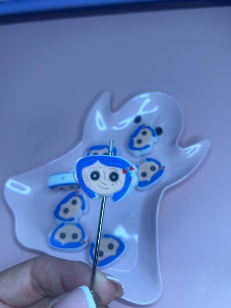 May include: A collection of blue and white character-themed charms and a decorative pick. The charms feature a cartoon face with blue hair and a red bow. The pick is held in a pink dish.