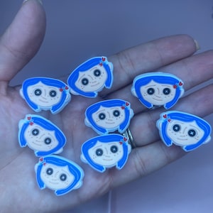 May include: A collection of cartoon character charms featuring blue hair, black button eyes, and a small red heart. The charms are white, blue, and tan. These charms are likely for shoes or accessories.