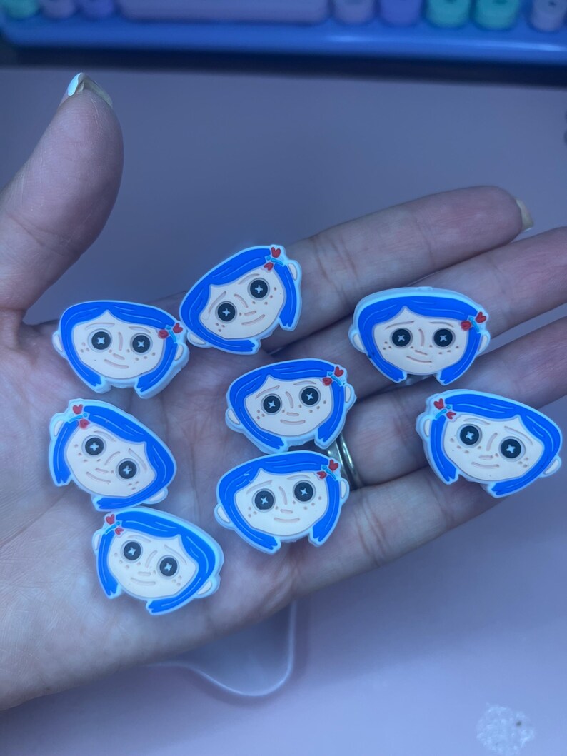 May include: A collection of cartoon character charms featuring blue hair, black button eyes, and a small red heart. The charms are white, blue, and tan. These charms are likely for shoes or accessories.