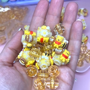 May include: A collection of yellow and clear beads, some featuring a cartoon bear character with red and yellow clothing. The beads are round and have a honey-themed design. Some beads have the word "HUNNY" on them.