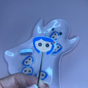 May include: A silver-toned pin featuring a cartoon character with blue hair, a red bow, and black eyes. The pin is held in front of a pink, ghost-shaped dish containing similar character-themed pins. The background is a solid pink.