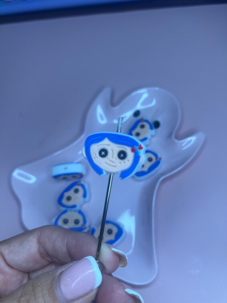 May include: A silver-toned pin featuring a cartoon character with blue hair, a red bow, and black eyes. The pin is held in front of a pink, ghost-shaped dish containing similar character-themed pins. The background is a solid pink.