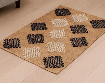 May include: A rectangular jute rug with a tan background and a repeating pattern of black and off-white squares. The rug is placed on a light-colored wooden floor, with a gray sofa and gold side table in the background.