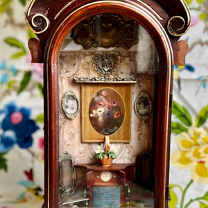 May include: A miniature wooden display case with a dark brown finish and gold accents. Inside, a detailed diorama features a small painting of flowers, miniature furniture, and framed portraits. The case has an arched top and a decorative base.