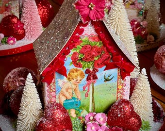 May include: A decorative miniature house with a glittered roof and a floral design. The house features a picture of a cherub watering a rose bush, surrounded by red bows, pink flowers, and glittered hearts. White bottle brush trees and red ornaments complete the festive scene.