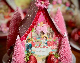 May include: A decorative pink miniature house with a glittered roof and a floral embellishment. The house features a vintage-style illustration with the text "To my Love". Pink bottle brush trees and heart-shaped decorations surround the house.