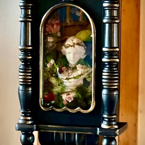 Puede incluir: Un mueble expositor de pared azul verdoso oscuro con detalles dorados. El mueble cuenta con una ventana central arqueada que muestra un busto blanco rodeado de vegetación y flores. El marco y los elementos decorativos están pintados en negro y dorado.