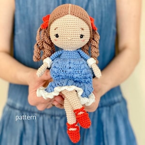 Puede incluir: Una muñeca de crochet con cabello castaño, un vestido azul y zapatos rojos. La muñeca está sentada en el regazo de una persona.